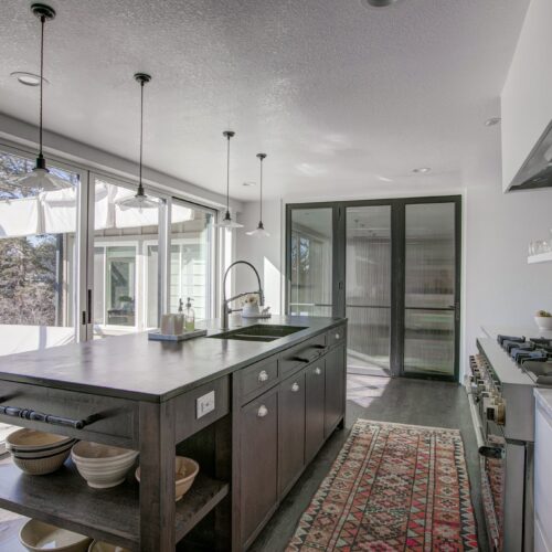 Modern kitchen design featuring a central island with dark cabinetry, stainless steel sink, and decorative rug, showcasing a bright and airy space with large windows and contemporary fixtures, reflecting BKC Kitchen and Bath's innovative remodeling style.