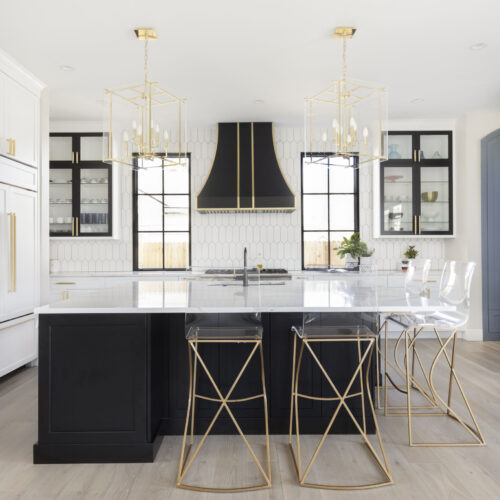 Black and white kitchen featuring a central island with a white marble countertop, gold-accented bar stools, modern pendant lighting, and a stylish black hood above the stove, reflecting contemporary design elements.
