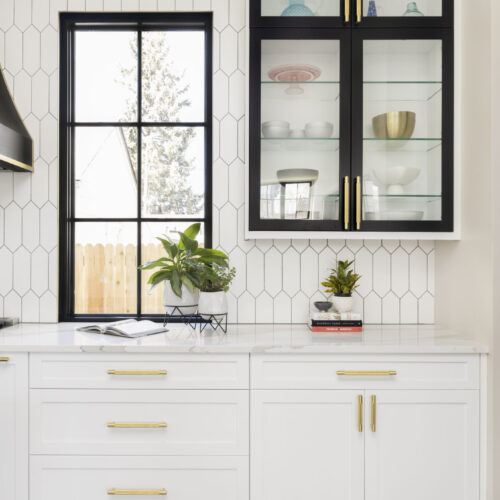 Modern kitchen featuring white cabinetry with gold hardware, black glass-front upper cabinets, and a geometric tile backsplash, complemented by a window with a view of greenery.