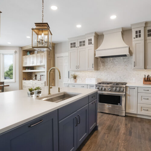 Transitional kitchen featuring gray and navy cabinetry, stainless steel appliances, and a large island with a farmhouse sink, illuminated by a pendant light.