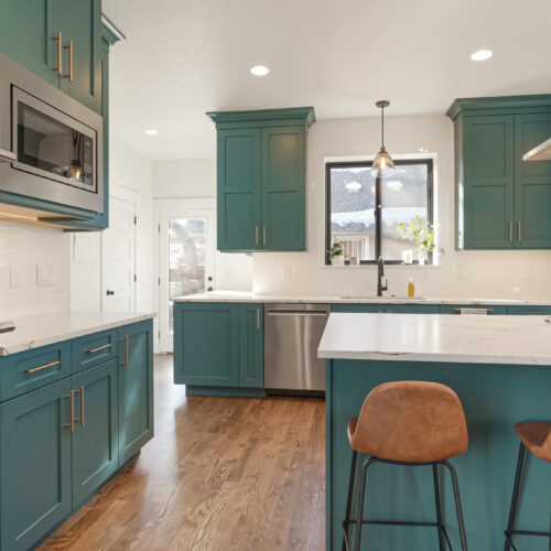 Teal cabinetry with white countertops and under-cabinet lighting in a modern kitchen setting, featuring a large window and wooden flooring.