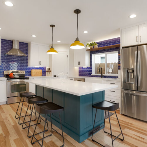 North Raleigh blue kitchen featuring white cabinetry, a large island with a teal base, modern pendant lighting, stainless steel appliances, and vibrant blue tile backsplash.