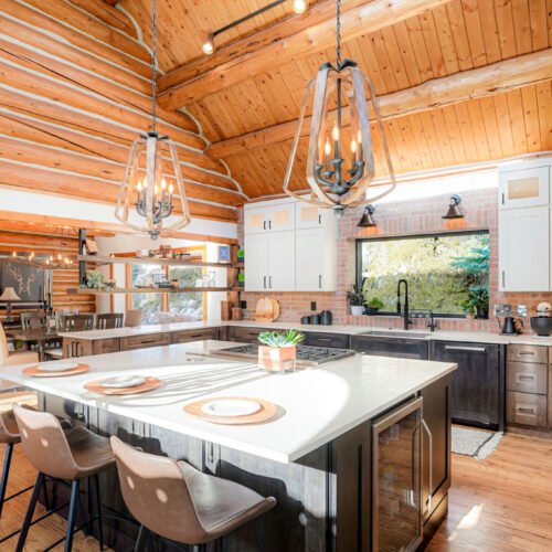 Modern kitchen design featuring white shaker cabinets, underlit countertops, and rustic wooden beams, with a large island and stylish pendant lighting, showcasing a blend of contemporary and mountain aesthetics.