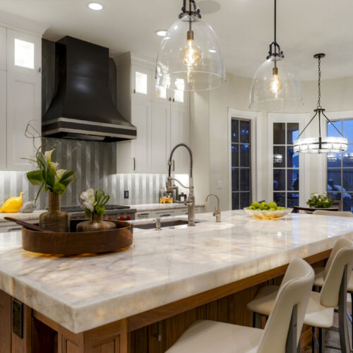 Modern kitchen featuring white shaker cabinets, underlit marble countertops, decorative pendant lighting, and a sleek black hood.